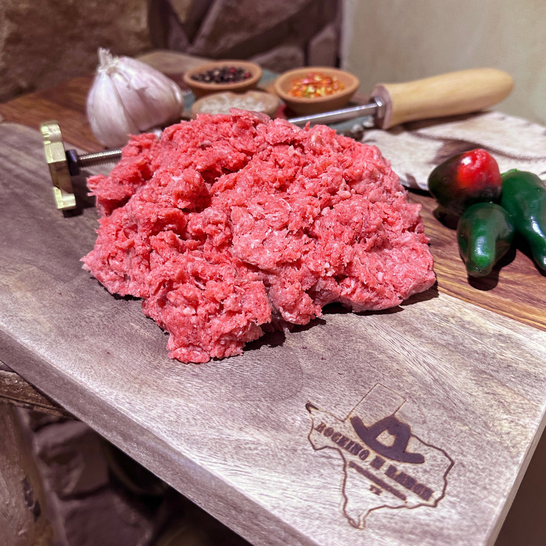 A pile of fresh ground beef on a wooden cutting board with spices, garlic, and green chili peppers in the background.