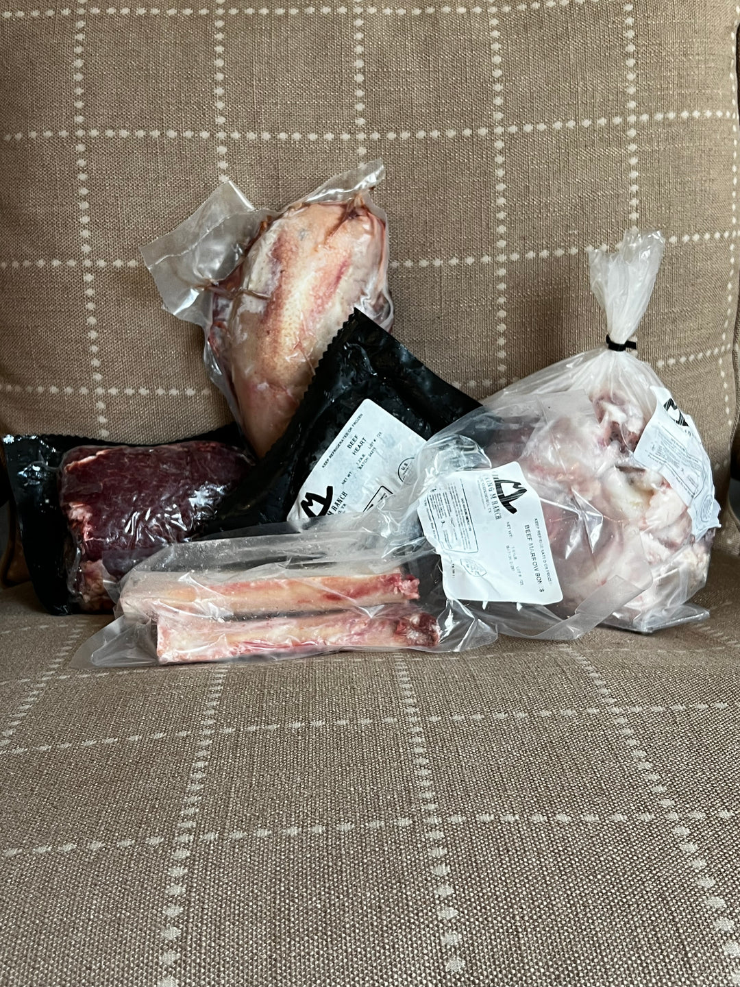 Assorted packages of meat including beef tongue, cheeks, oxtail, knuckle bones, and marrow bones, placed on a sofa cushion.