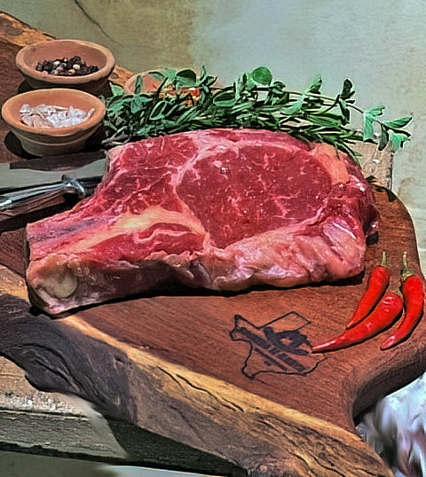 A bone-in rib eye steak on a wooden cutting board, accompanied by seasonings, herbs, and decorative red chili peppers.